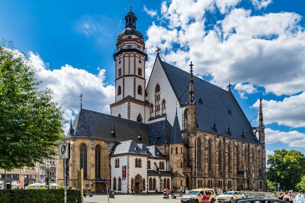 Die Thomaskirche in Leipzig befindet sich in der Mitte des Bildes. Vor der Kirche laufen Menschen und Autos parken. Neben der Kirche sind Bäume.