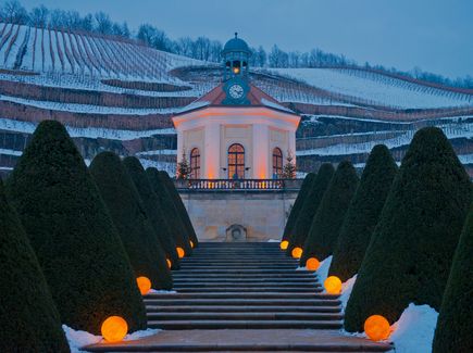 Das Schloss Wackerbarth im Winter ist abgebildet. Im Hintergrund sind Weinberg und alles ist mit Schnee bedeckt. Es ist abends und Lichter leuchten. 