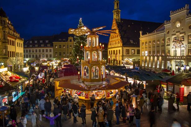 Ein Marktplatz ist weihnachtlich geschmückt. In seiner Mitte steht eine große Weihnachtspyramide. Verschiedene Stände und Buden säumen den Platz. Es sind zahlreiche Menschen unterwegs.