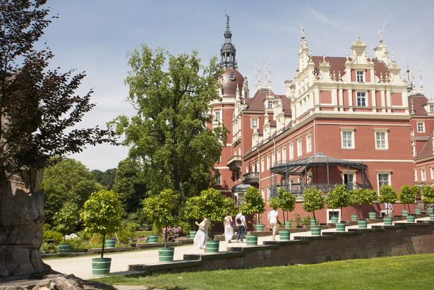 Schloss Bad Muskau im Fürst-Pückler Park ist auf dem Bild. Das Schloss ist auf der rechten Seite und auf der linken Seite sind vereinzelte Bäume und ein Weg, der zum Schloss führt. Auf dem Weg stehen Töpfe mit kleinen Bäume und vier Menschen laufen auf dem Weg.