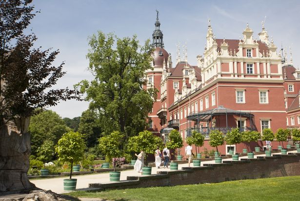 Schloss Bad Muskau im Fürst-Pückler Park ist auf dem Bild. Das Schloss ist auf der rechten Seite und auf der linken Seite sind vereinzelte Bäume und ein Weg, der zum Schloss führt. Auf dem Weg stehen Töpfe mit kleinen Bäume und vier Menschen laufen auf dem Weg.