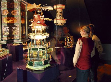 Zwei Besucher schauen sich eine Pyramide und Holzkunst in der Manufaktur der Träume in Annaberg Buchholz an. Der Raum ist dunkel und die Ausstellungsstücke leuchten.