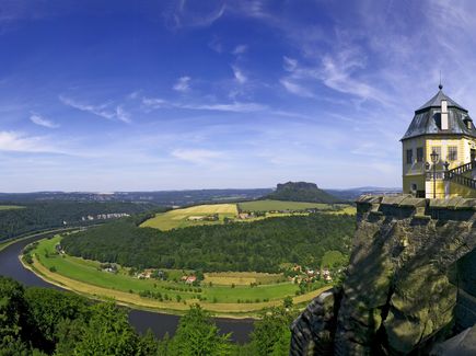 In dem Bild ist der Blick von der Festung Königsstein auf die Sächsische Schweiz zu sehen. Neben der Festung fließt der Fluss Elbe durch die Landschaft der Sächsischen Schweiz. Die Landschaft besteht aus Wälder, Feldern, Wiesen, vereinzelten Häusern und in der Ferne befinden sich Felsen.