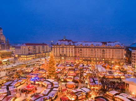 Der Dresdner Striezelmarkt ist abgebildet. Es ist dunkel und alle Lichter leuchten. Auf dem Markt stehen viele Stände. Links neben dem Markt ist eine große Straße. Neben dem Markt stehen Häuser. Es hat geschneit und die Dächer sind schneebedeckt.