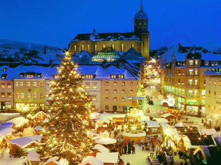 Der Weihnachtsmarkt in Annaberg Buchholz ist von oben zu sehen. In der Mitte des Marktes steht ein großer Weihnachtsbaum und daneben eine Pyramide. Um den Markt herum stehen Häuser und im Hintergrund eine große Kirche. Alles ist schneebedeckt und alle Lichter leuchten.