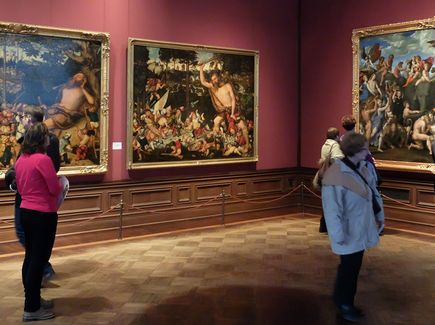 Ein Raum der Gemäldegalerie "Alte Meister" in Dresden ist abgebildet. In dem Raum hängen drei große Bilder. Besucher schauen sich diese Bilder an.