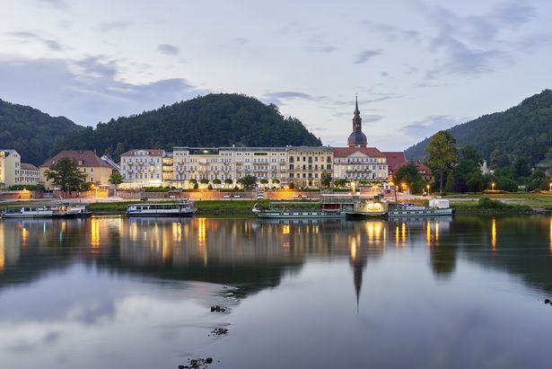 Das Hotel Elbresidenz Bad Schandau ist von außen abgebildet. Vor dem Hotel ist der Fluss Elbe, auf dem Schiffe liegen. Es ist abends und Lichter leuchten.