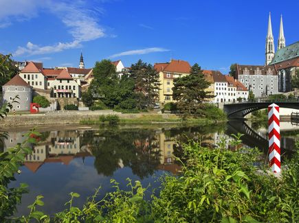 Der Fluss Neiße und das Panorama von Görlitz sind zu sehen. Über den Fluss geht ein Brücke. Das Panorama besteht aus Häusern vor denen ein paar Bäume stehen.