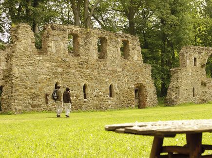 Auf einer Wiese des Kloster Nimbschen laufen zwei Leute neben einer alten Mauer. Eine Bank mit Tisch steht auf der Wiese.