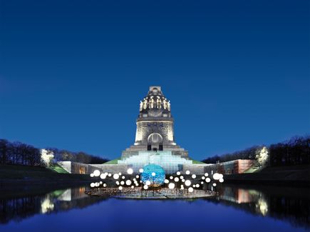 Das Völkerschlachtdenkmal in Leipzig ist abgebildet. Es ist nachts und das Denkmal ist angeleuchtet sowie befinden sich Lichtinstallationen vor dem Denkmal.