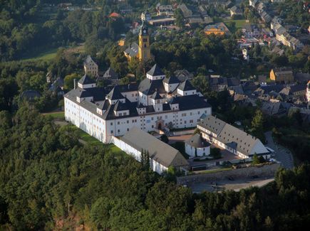 Die Augustusburg ist von oben zu sehen. Um die Burg herum befindet sich ein Wald und rechts neben der Burg Häuser.
