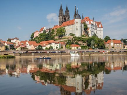 Die Albrechtsburg in Meißen ist abgebildet. Um die Burg herum stehen Häuser. Vor den Häusern fließt der Fluss Elbe. Die Albrechtsburg und die Häuser spiegeln sich in dem Fluss. 