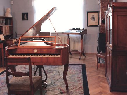 Ein Raum im Schumann Haus in Zwickau ist abgebildet. In der Mitte des Raums steht ein alter Flügel und ein Stuhl auf einem Teppich. An den Wänden stehen alte Möbel und der Raum hat zwei Fenster.