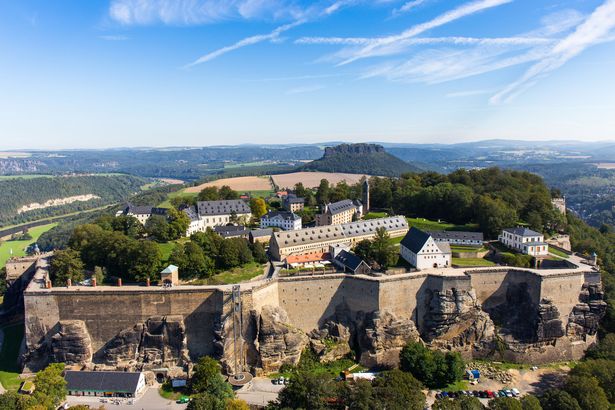 Die Festung Königstein ist von oben zu sehen. Die Festung wird von einer Mauer umrahmt. Außerhalb der Mauer sind Wiese und Wälder. Im Hintergrund der Festung befindet sich der Lilienstein.