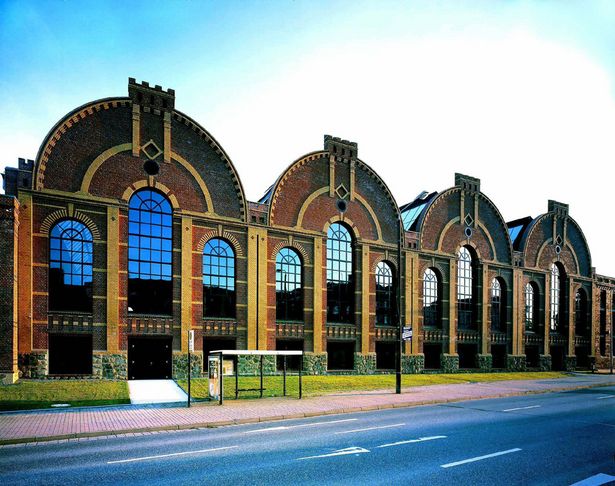 Auf dem Bild ist das Sächsische Industriemuseum in Chemnitz von außen zu sehen. Das Gebäude hat viele große Fenster und besteht aus roten Steinen. Vor dem Museum ist eine Straße, eine Bushaltestelle und ein Stück Wiese.