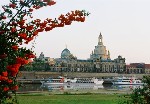 Das Dresdner Elbufer mit der Frauenkirche ist auf dem Bild. Auf der Elbe sind zwei Schiffe, auf der linken Seite des Bildes ein Baum und im Vordergrund eine Wiese. 