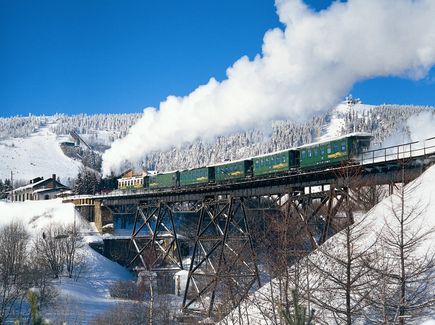 Die Fichtelbergbahn im Erzgebirge ist abgebildet. Sie fährt über eine Brücke in einen Bahnhof ein, der sich in einer schneebedeckten Landschaft befindet. Im Hintergrund ist ein Berg mit Bäumen.