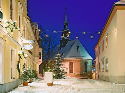 Die Bergkirche in Annaberg Buchholz ist im Winter zu sehen. Vor der Kirche und auf dem Dach der Kirche liegt Schnee. Neben der Kirche stehen Häuser. Es ist Nacht und Lichter leuchten.