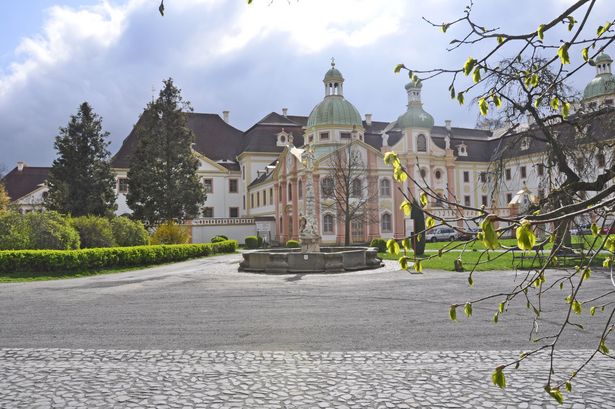 Auf dem Bild ist das Kloster St. Marienthal in Ostriz abgebildet. Vor dem Kloster ist ein Weg und ein Brunnen sowie eine Hecke, Wiese und Bäume.