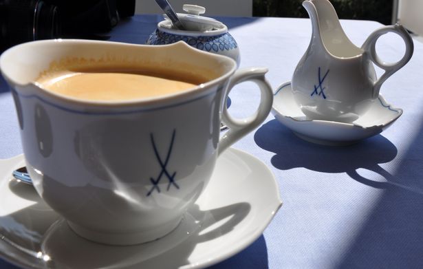 Eine Kaffeetasse aus Meissner Porzellan steht auf einem kleine Teller. Auf der Tasse ist das Symbol für das Meissner Porzellan - zwei sich kreuzende Schwerter abgebildet. Im Hintergrund ist eine kleine Kanne mit Milch ebenfalls aus Meissner Porzellan. Beides steht auf einem Tisch.